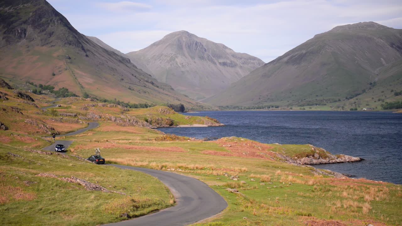 People driving on a road by the lake towards the mountains of the England Lake District - wide shot