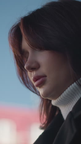 mujer con cabello marrón suave y expresión reflexiva en un entorno al aire libre, con abrigo negro y galleta blanca, contra un fondo urbano borroso y un cielo azul claro