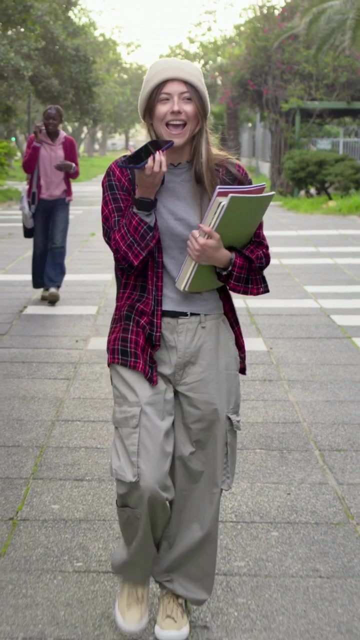 Video verticale: Donna che cammina con un berretto, tiene dei quaderni e parla al telefono su un sentiero del campus.