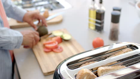 Slicing avocado and tomato, man preparing breakfast with toaster in kitchen
