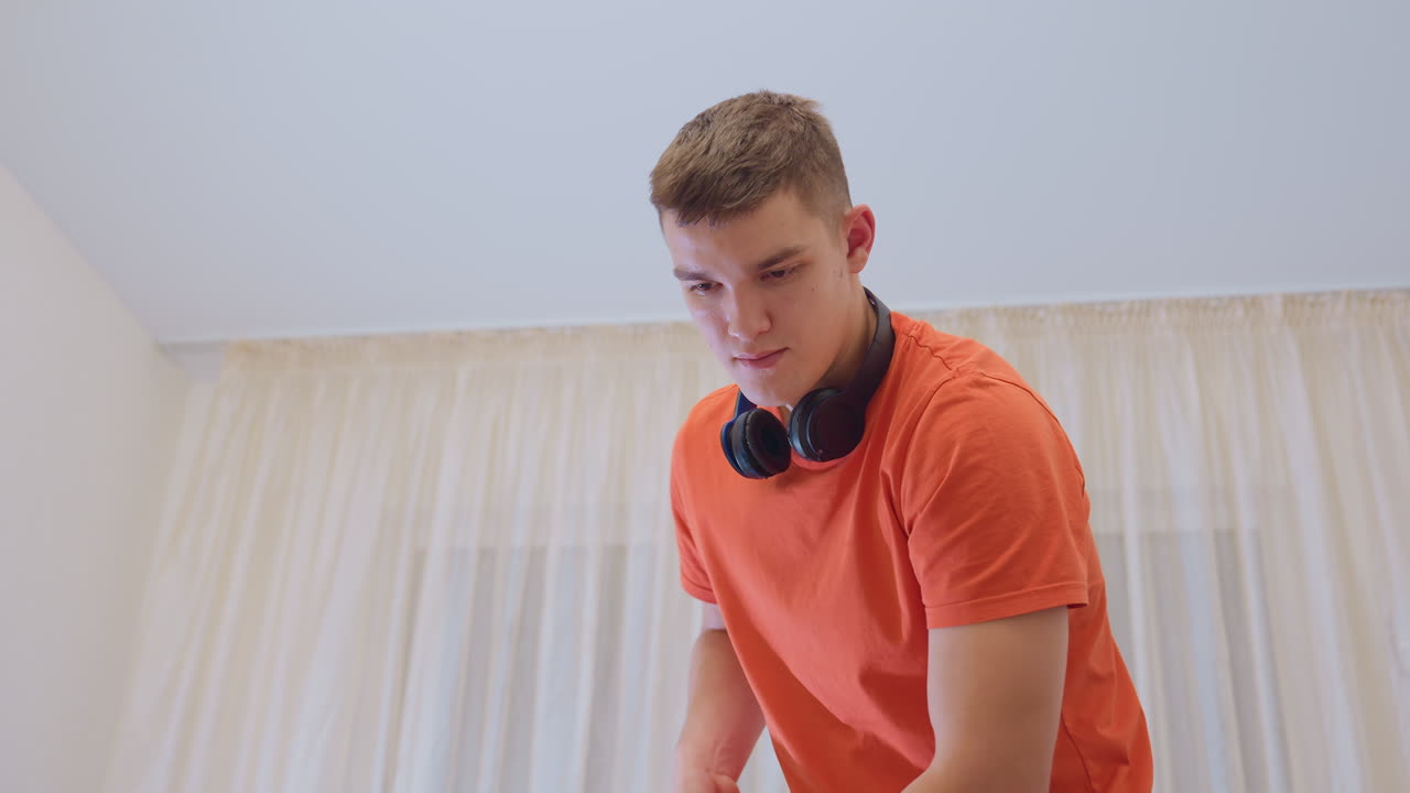 Young man in orange shirt with headphones around neck bends forward while mopping floor indoors near curtains showing focus on household cleaning hygiene and daily domestic routine