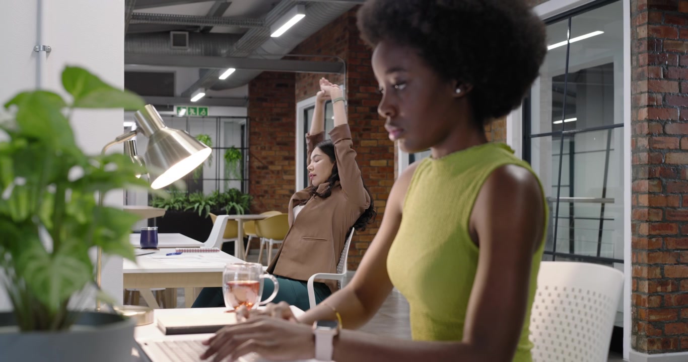 Female coworkers, foreground woman typing on laptop while colleague stretching after reading papers