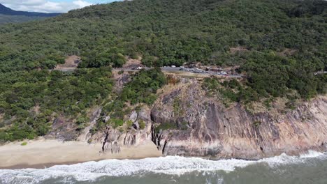 4k aerial view of rocky cliff and a small sandy beach with a coastal highway in the background