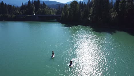 aerial reversing over stand up paddle boarders, floating on an emerald lake, paddling away from shore next to a highway surrounded by trees, forest mountains, islands on a bright blue sunny day