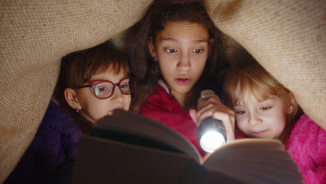 children girl kids under blanket reading interesting fairytale story book using flashlight at home