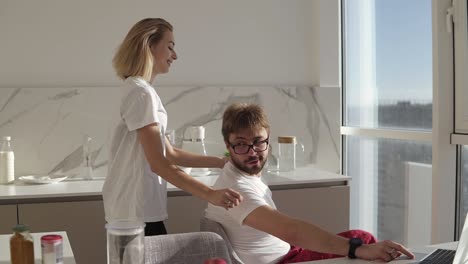 bearded man in eyes glasses working on laptop computer sitting at home on a kitchen with panoramic windows. young wife embracing him from behind and they kissing each other