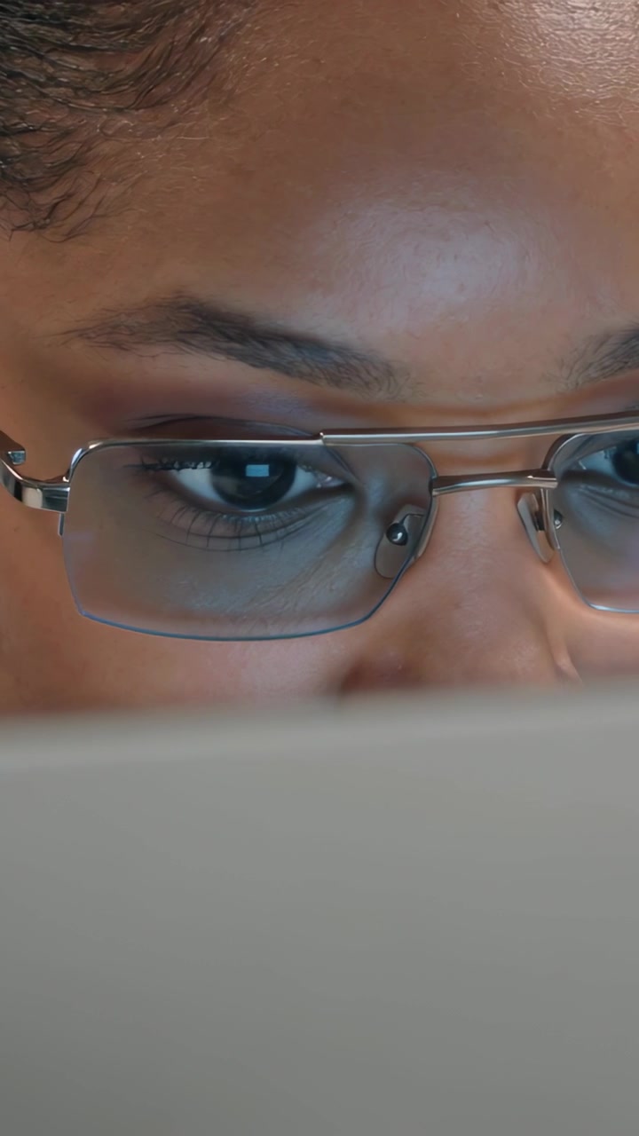 Video verticale: Donna con gli occhiali che studia, guarda oltre il bordo del laptop alla scrivania, con un riflesso della lente.