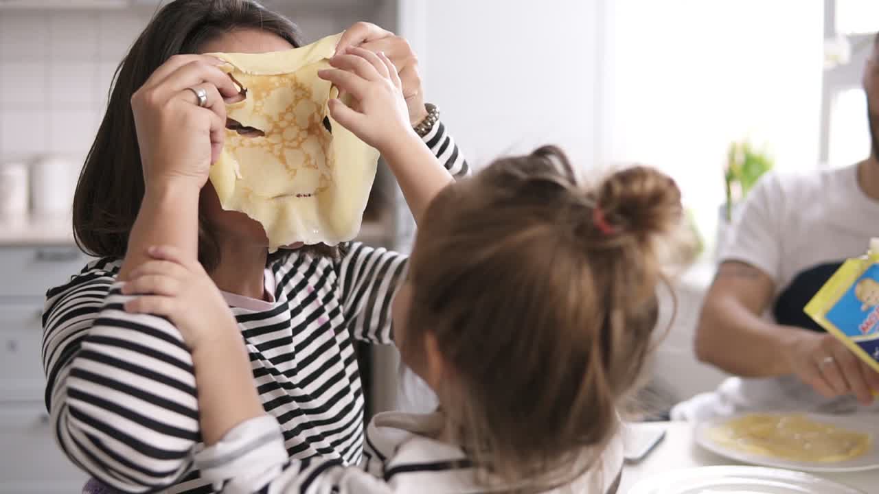 Funny mother is playing with her little daughter making comical mask with pancake on her face. Family breakfast