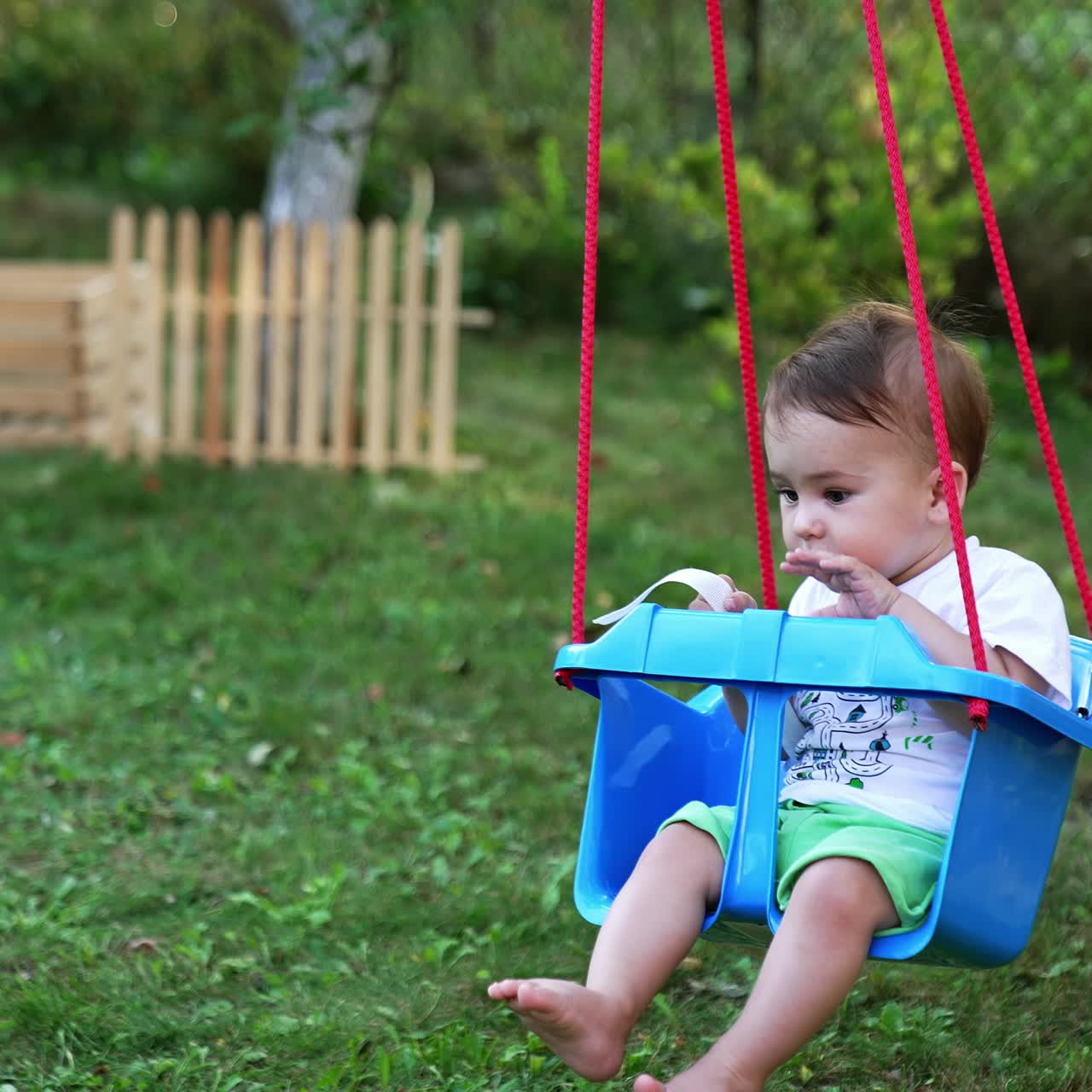 Peaceful Caucasian baby boy swaying in the blue swing in the garden. Trees and wooden fence at backdrop