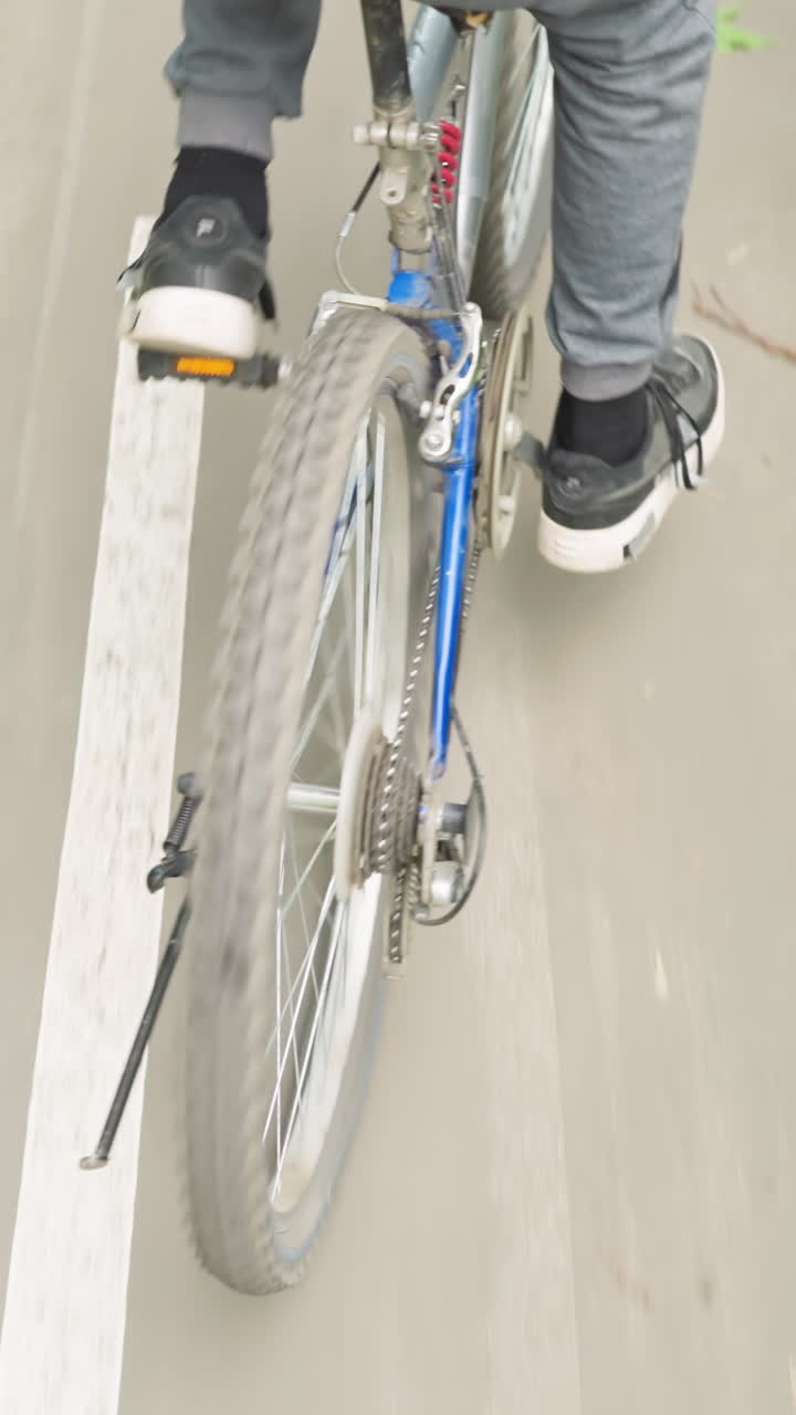 Back view of someone cycling along a wet paved road with white markings, surrounded by lush roadside greenery, the cyclist pedals off, leaving water tracks on the road surface