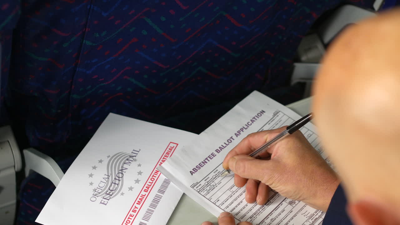 A bald man filling out an absentee ballot application with a pen while seated on an airplane. The "Official Election Mail" envelope is placed nearby for mail-in voting while traveling
