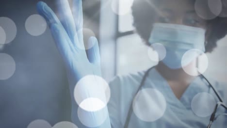 animation of glowing light spots over african american female doctor wearing medical gloves