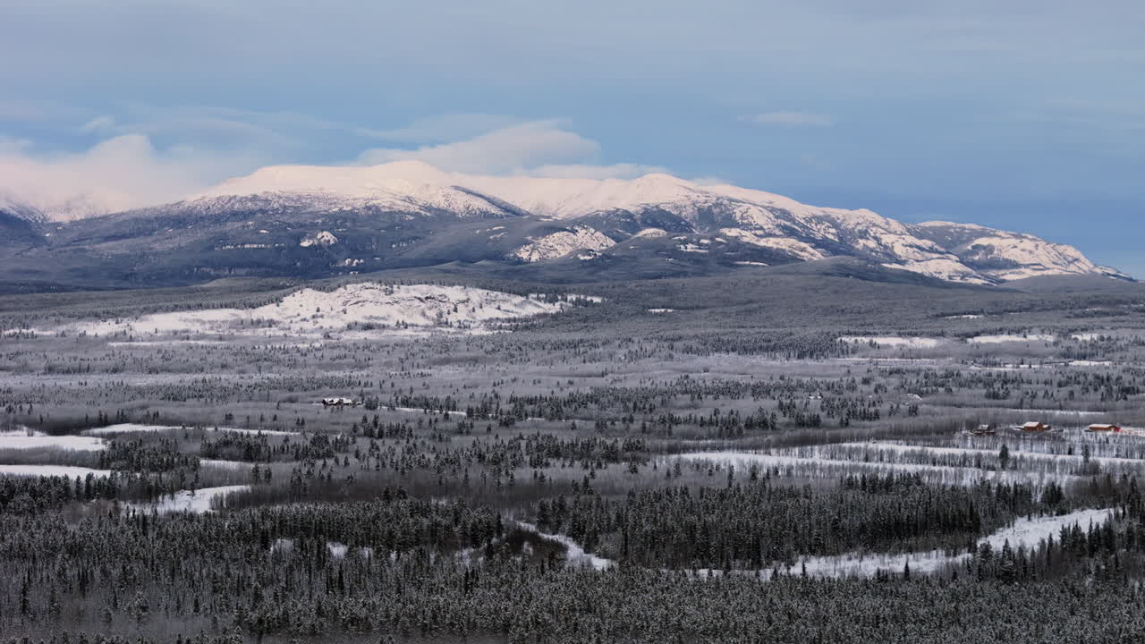 This cinematic aerial captures a snowy sunset over Ibex Valley in Yukon. Mountains and fir forests draped in winter white evoke the vastness and isolation of the northern wilderness.