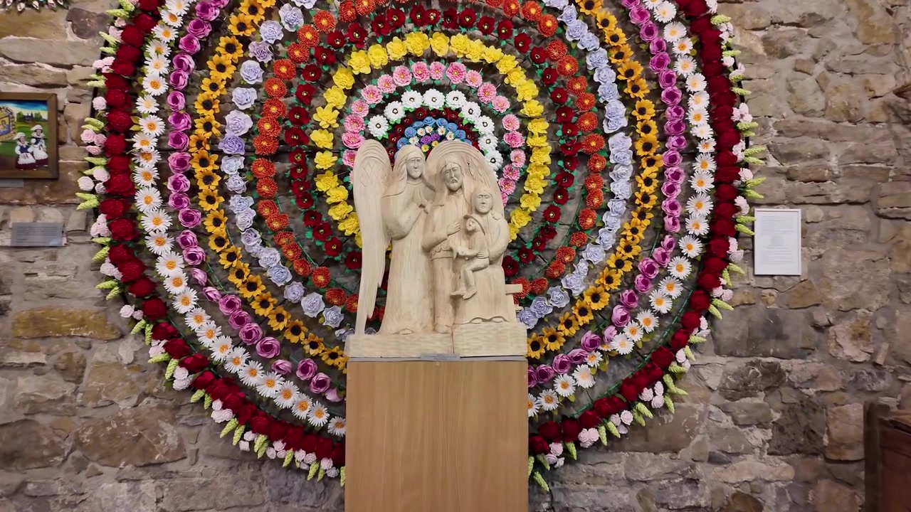 Anjo de madeira lindamente esculpido com figuras de família, apresentado diante de uma vibrante decoração floral circular em um ambiente de museu.