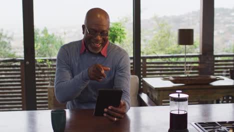 Happy-african-american-senior-man-sitting-at-counter-in-kitchen-making-video-call-with-smartphone