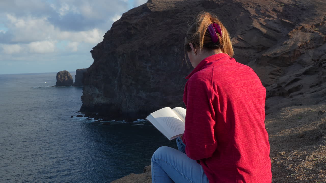 Woman enjoys reading a book on a cliff overlooking Roque Partido and the atlantic ocean in Gran Canaria, canary islands. Slow motion
