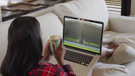 compuesto de una mujer sentada en casa sosteniendo un café viendo un evento de atletismo en una computadora portátil