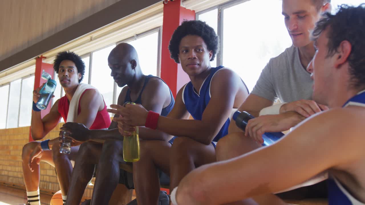 Diverse male basketball team and coach resting and drinking water after match