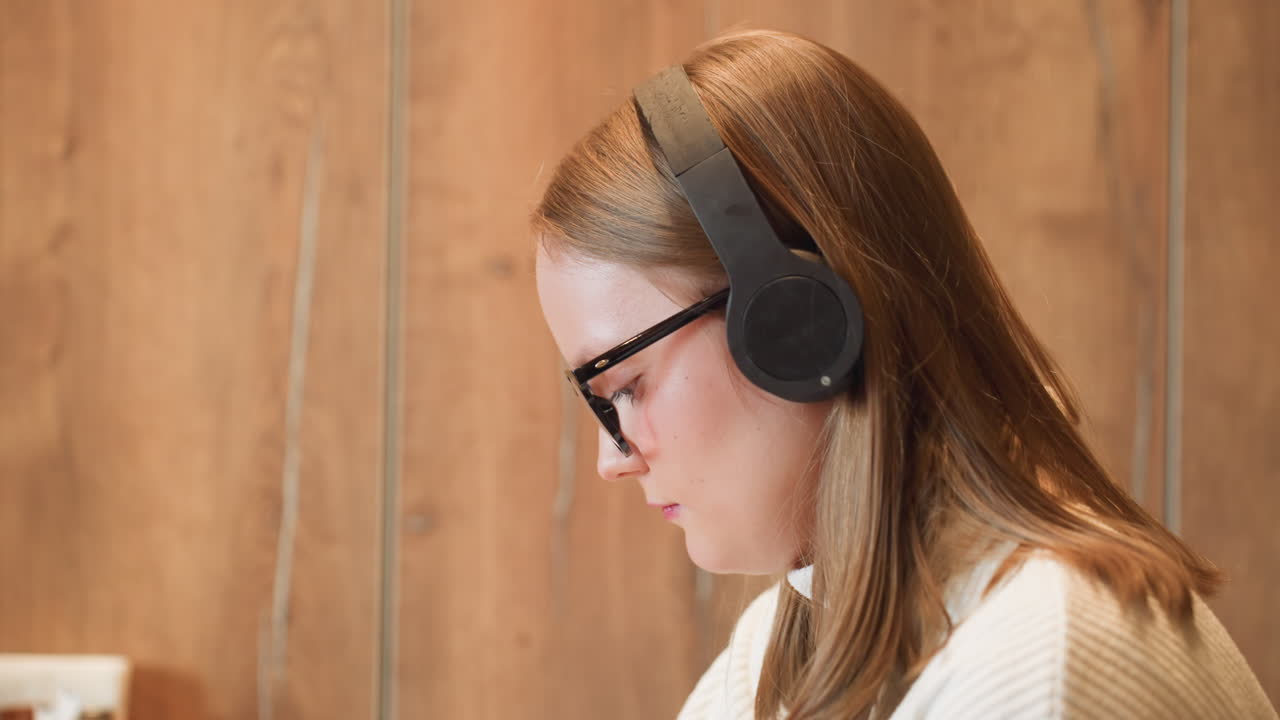 Side view close up of young woman in glasses and white sweater wearing headphone, gently nodding to music in relaxed mood, with warm wooden paneling in soft background