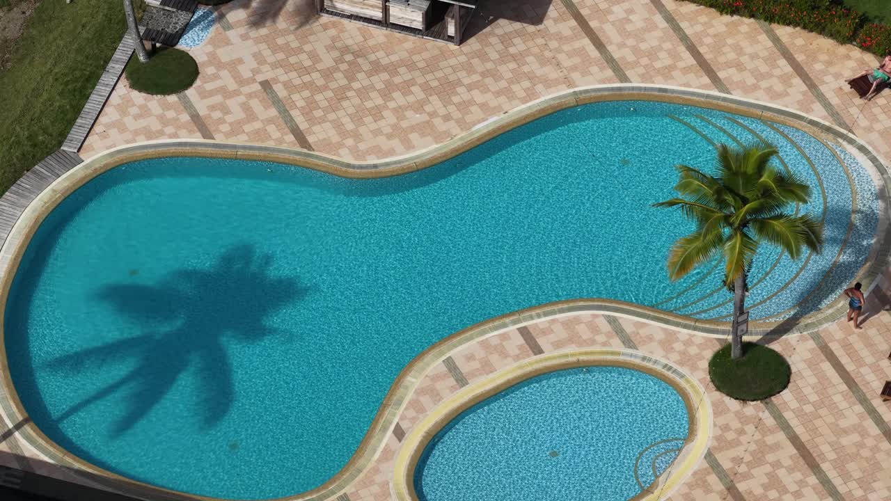 Højt vinkel luftfoto af en tropisk feriesteds swimmingpool med palmer og skygger.