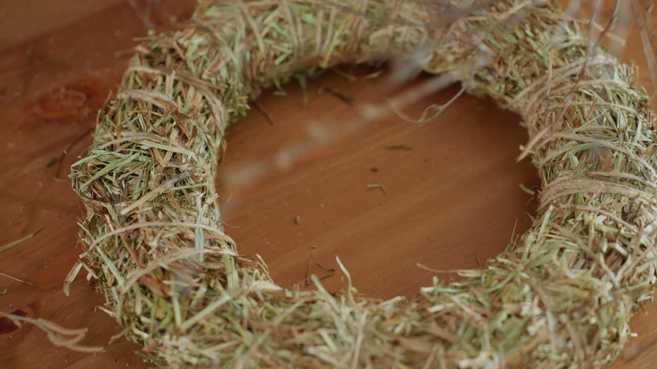 Female florist holding rustic twig wreath in hands while working on straw base, arranging branches with care during creative handmade craft process, showcasing artisan
