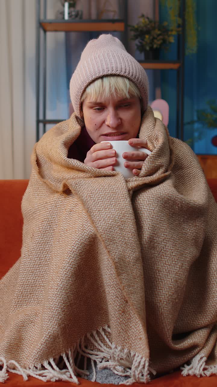 Una donna malata e malata indossa un cappello avvolto in un quadri si siede tremando di freddo sul divano a bere tè caldo