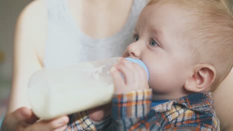 un niño encantador come comida de la botella en los brazos de la madre de cerca