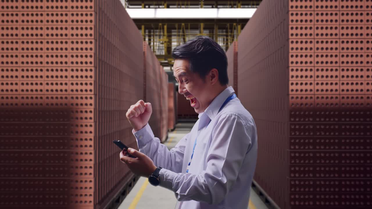Side View Of An Asian Male Professional Worker With His Smartphone With Red Brick Packed in Stacks Are Stored, He Raises His Fist Up With Screaming Goal After Check On The Smartphone