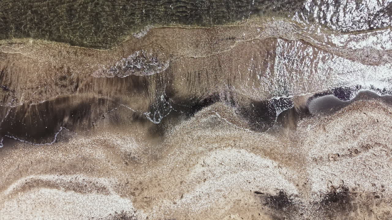 Le onde si infrangono sulla spiaggia sabbiosa con la bassa marea in una zona costiera, rivelando motivi di sabbia e acqua sotto il sole splendente.