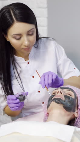 pretty cosmetologist with long hair makes a mask to a woman for cleansing the pores on the face in cosmetology. cosmetic procedures. spa vertical video
