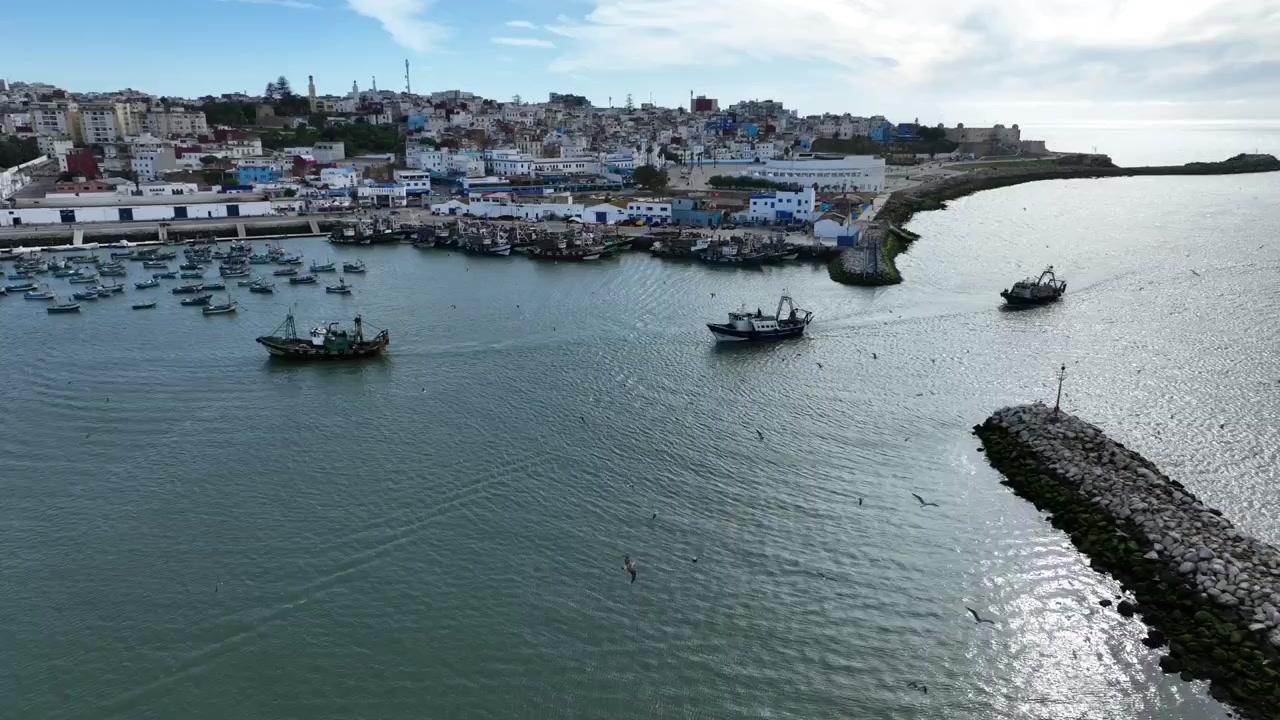Vista aerea di pescherecci che navigano un canale portuale verso l'Oceano Atlantico in Marocco, mostrando traffico navale attivo, frangiflutti e infrastrutture portuali operative