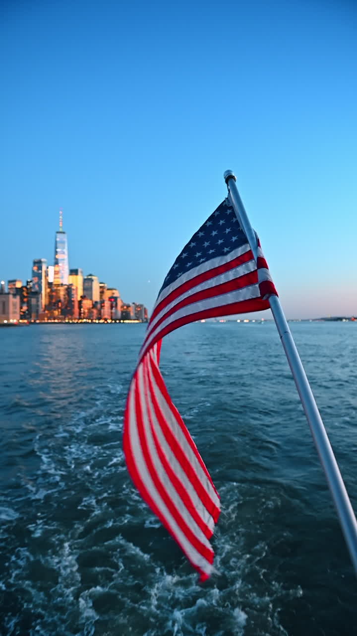 Bandiera americana che sventola su una barca con lo skyline di New York sullo sfondo. Una barca naviga sul fiume con la bandiera americana che sventola nel vento. Sullo sfondo si erge lo skyline di New York con il One World Trade Center visibile.