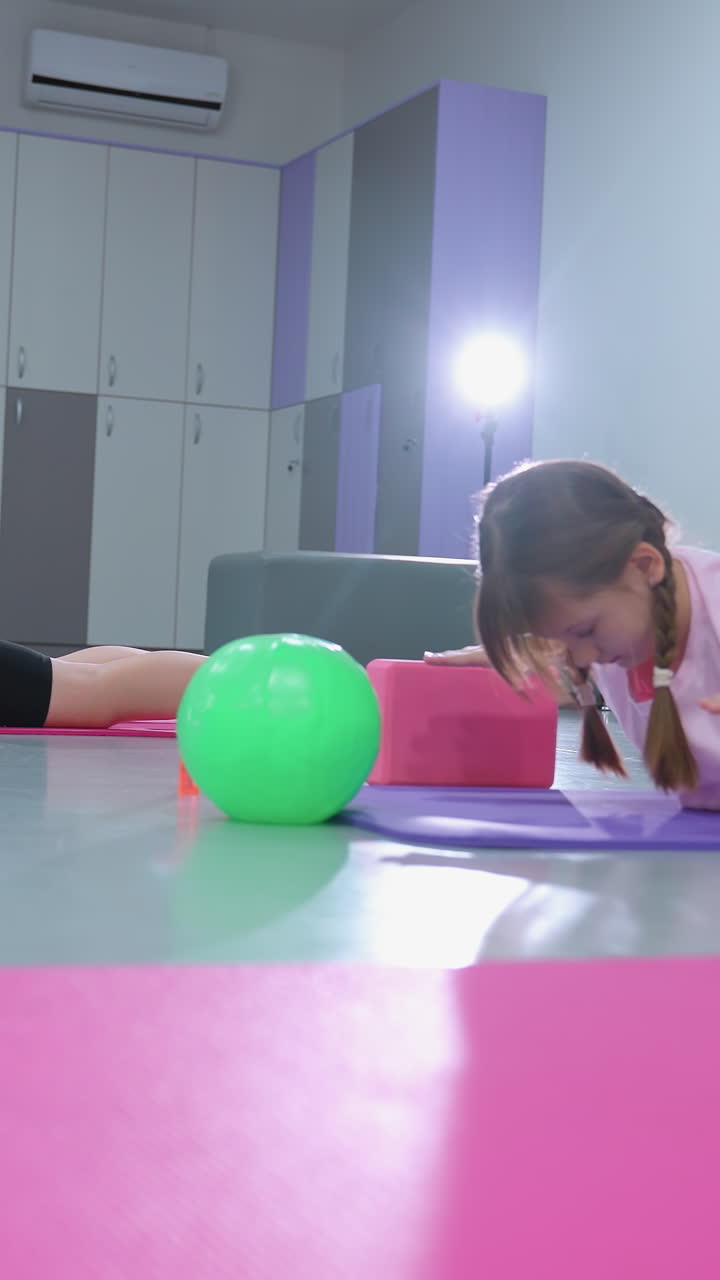 ids en una sesión de gimnasia ligeramente elevándose en esteras, estirándose hacia adelante, con el instructor parcialmente visible observando, en medio de coloridos equipos de gimnasio