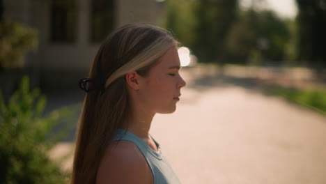 close-up of lady walking with focused look, blonde hair clipped back, sunlight reflecting on her, with blurred background featuring greenery and light reflections near building in afternoon light