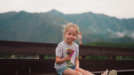 blonde child holds wild flowers sitting on wooden bench