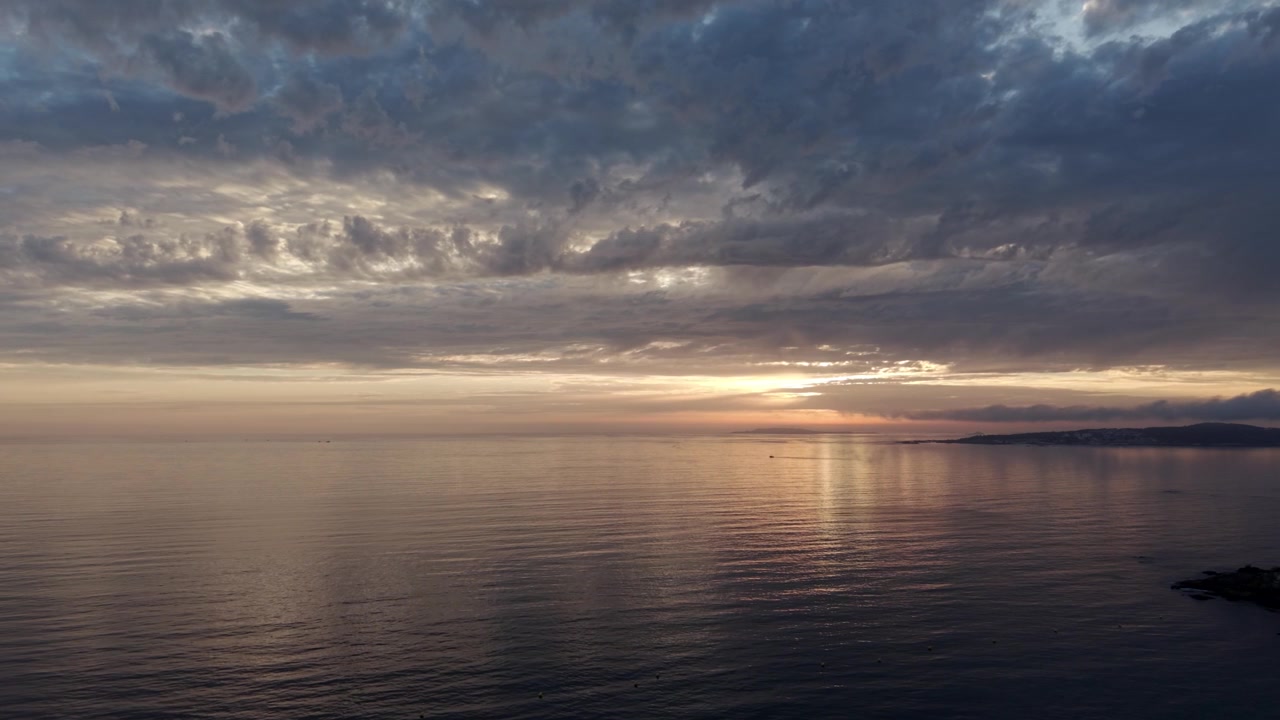 Aerial drone view of a soft moody sunset with muted golden light reflecting on very calm sea water under layered clouds on the coast of Galicia Spain