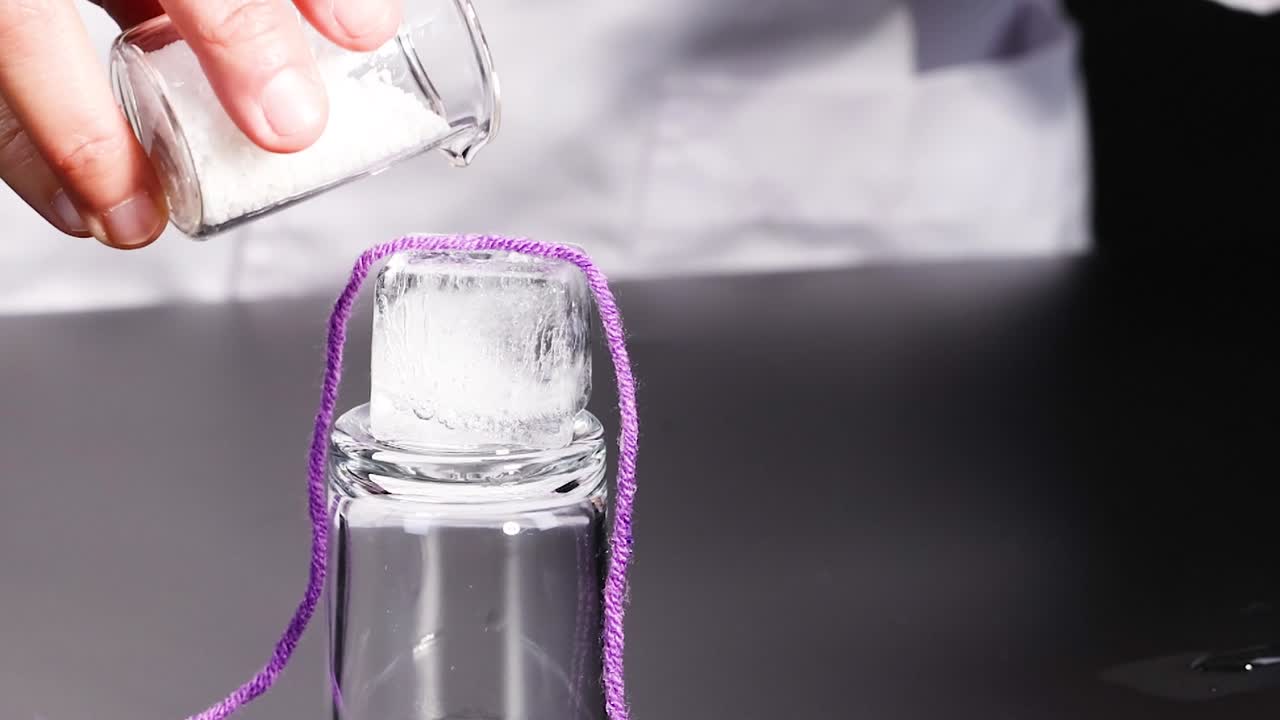 A close-up view of salt being poured onto an ice cube wrapped with a purple string.