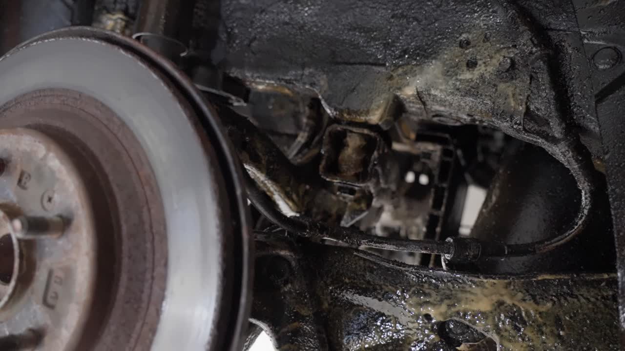 Handheld close-up looking past rear brake disc to suspension and chassis showing control arms, beams coated with lanolin rust protection product