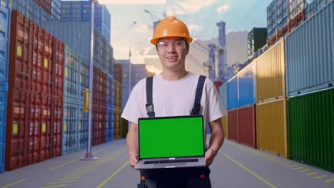 Asian Man Worker Wearing Goggles And Safety Helmet Smiling And Showing Green Screen Laptop To The Camera While Standing At Container Yard Warehouse
