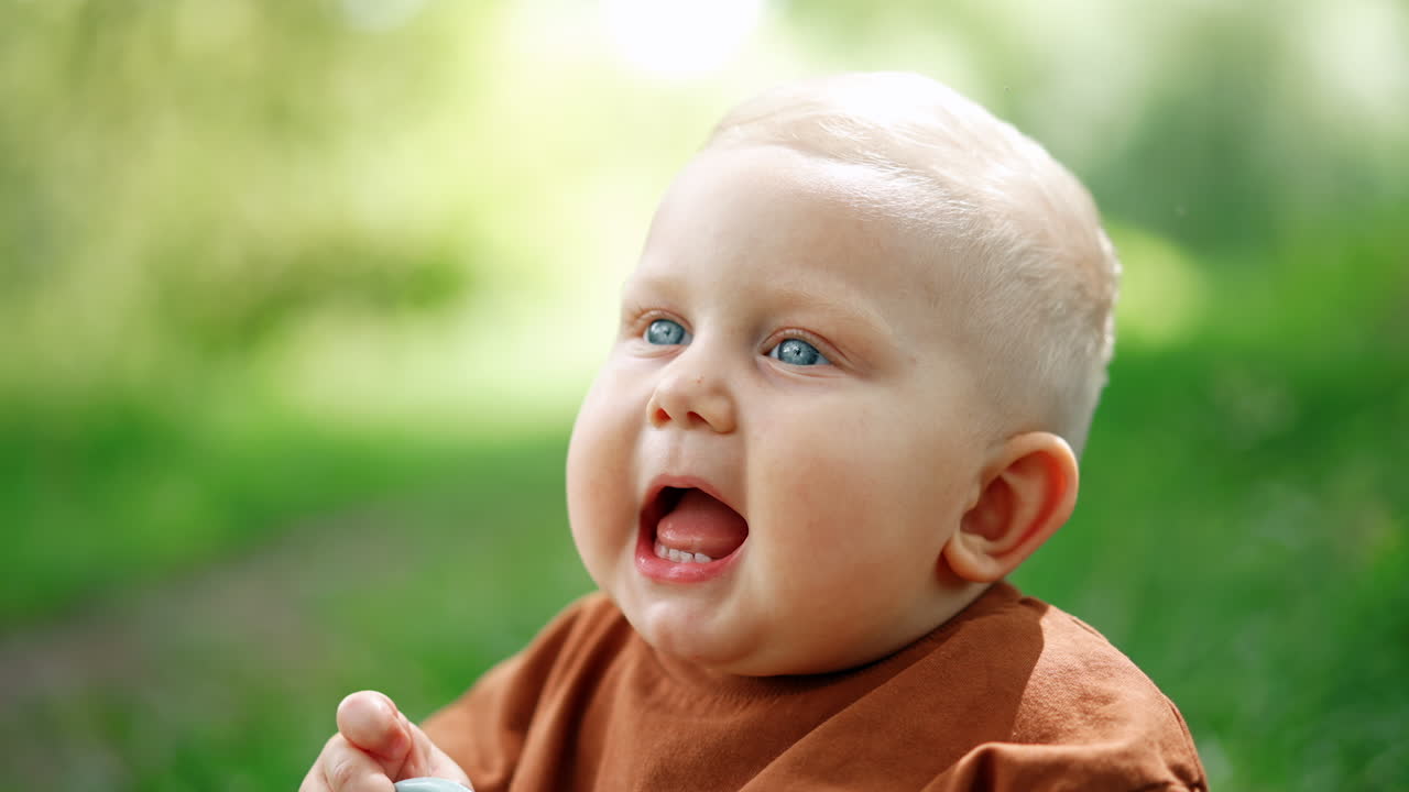 Adorable blond little baby boy with blue eyes and plump cheeks. Portrait of a lovely child sitting outdoors.