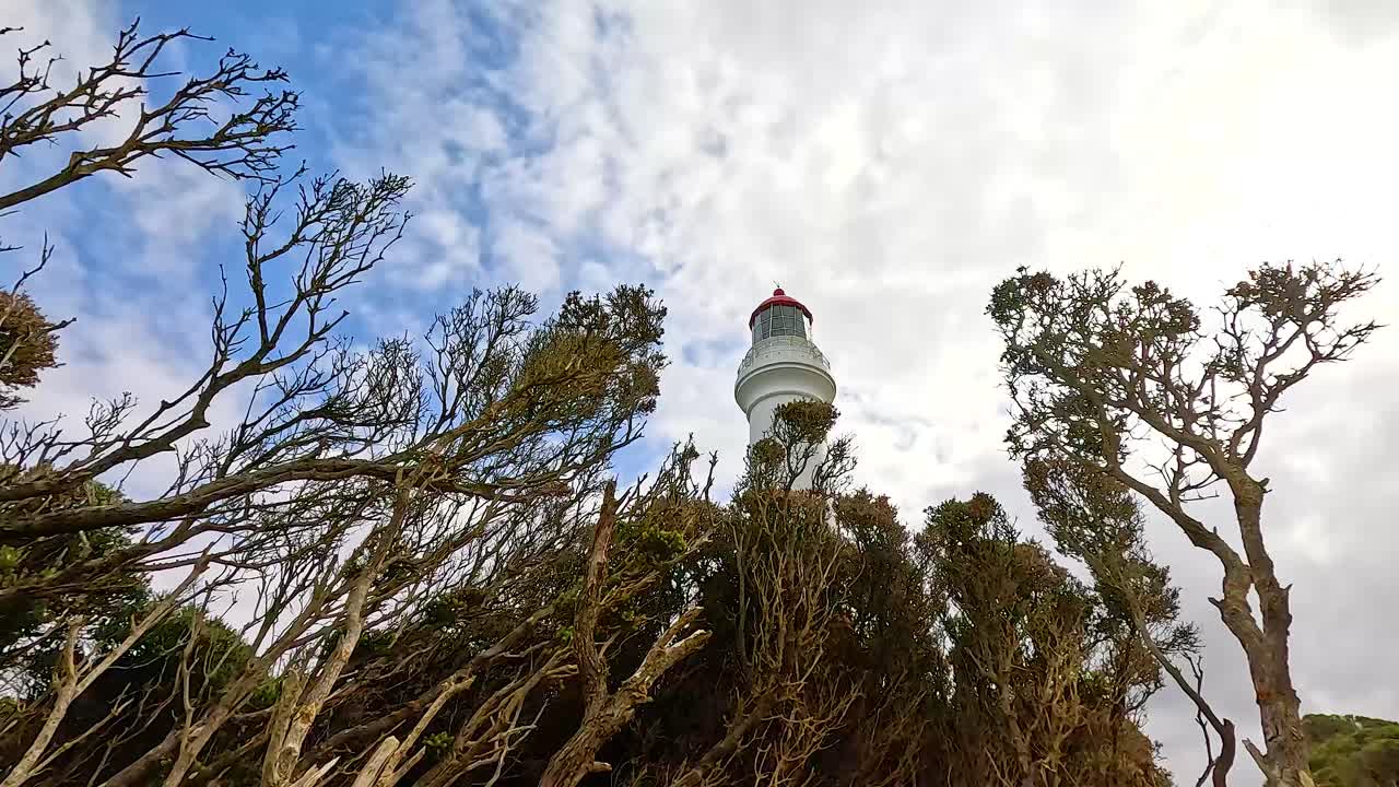 A lighthouse stands amidst windswept trees under a partly cloudy sky, capturing a serene coastal scene