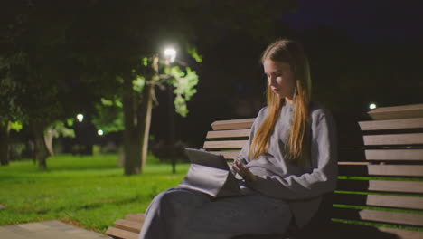 young girl sitting on park bench at night, fully immersed in her tablet under soft park lighting, people stroll in the blurred background