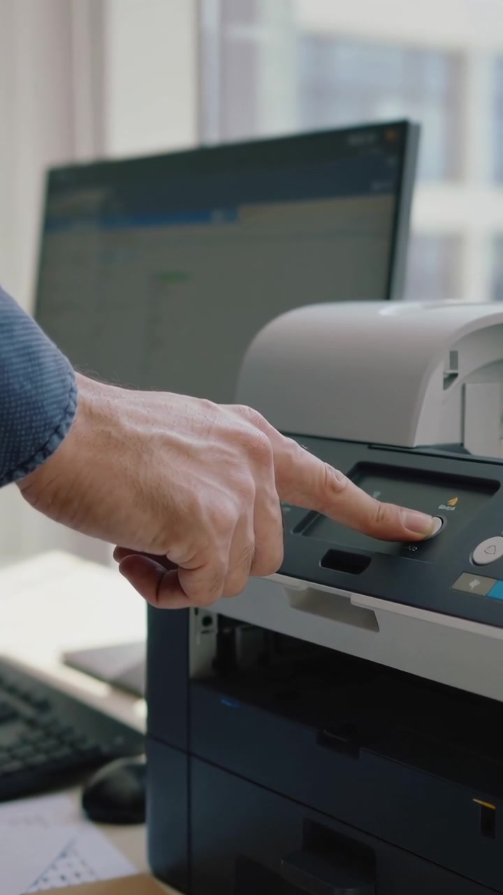 Vertical video: Hand entering frame in blue shirt tapping printer control at desk, starting print