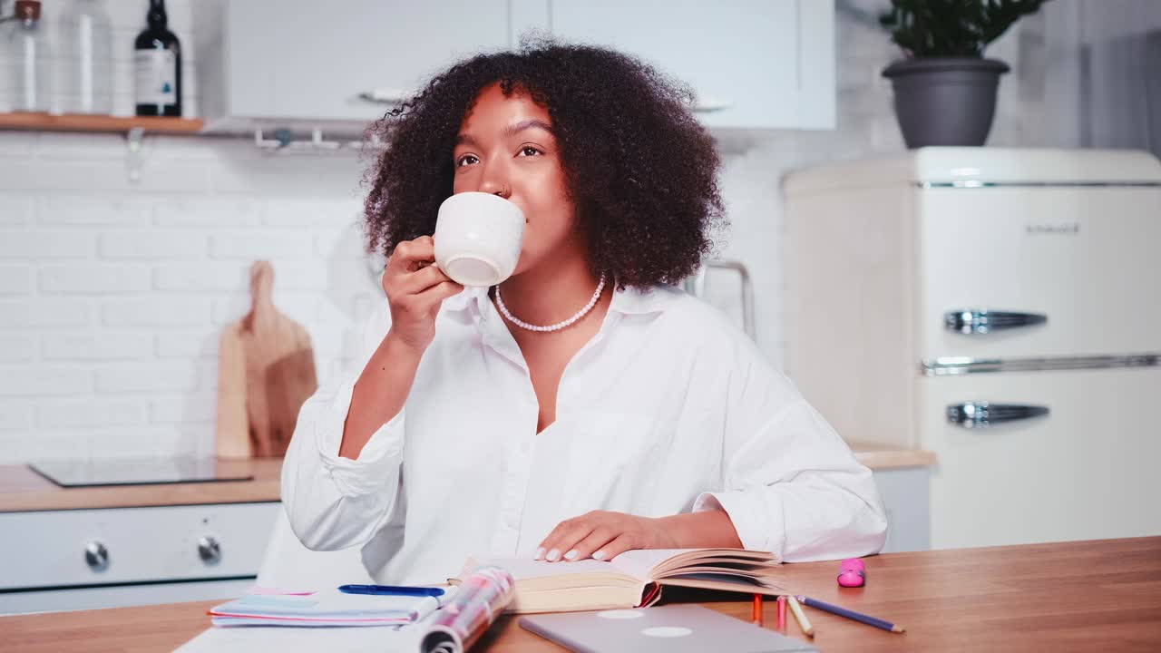 Young african american woman reads book and drinks coffee sits at kitchen