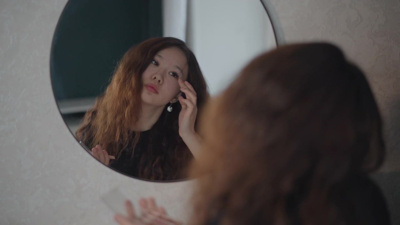 Donna con capelli ricci in una zona trucco del bagno illuminata delicatamente, sta davanti a uno specchio rotondo a parete, si sistema i capelli, si tocca il viso, si allaccia una collana e controlla il suo riflesso durante una tranquilla routine personale.