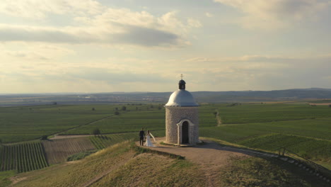 Una-Pareja-De-Recién-Casados-Caminando-Tomados-De-La-Mano-Alrededor-De-Una-Pequeña-Capilla-En-Una-Colina-Con-Vista-A-Los-Viñedos,-órbita-Aérea