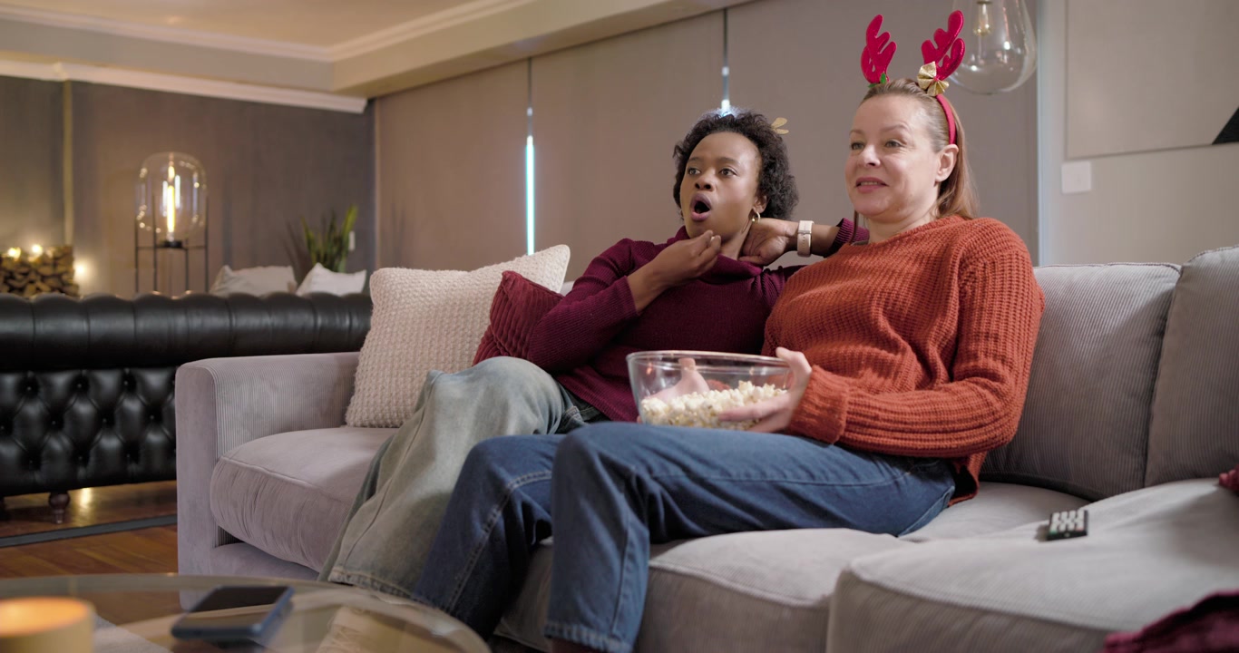 Donne diverse sedute sul divano a casa, che premono il telecomando della TV, gestendo il programma e mangiando popcorn