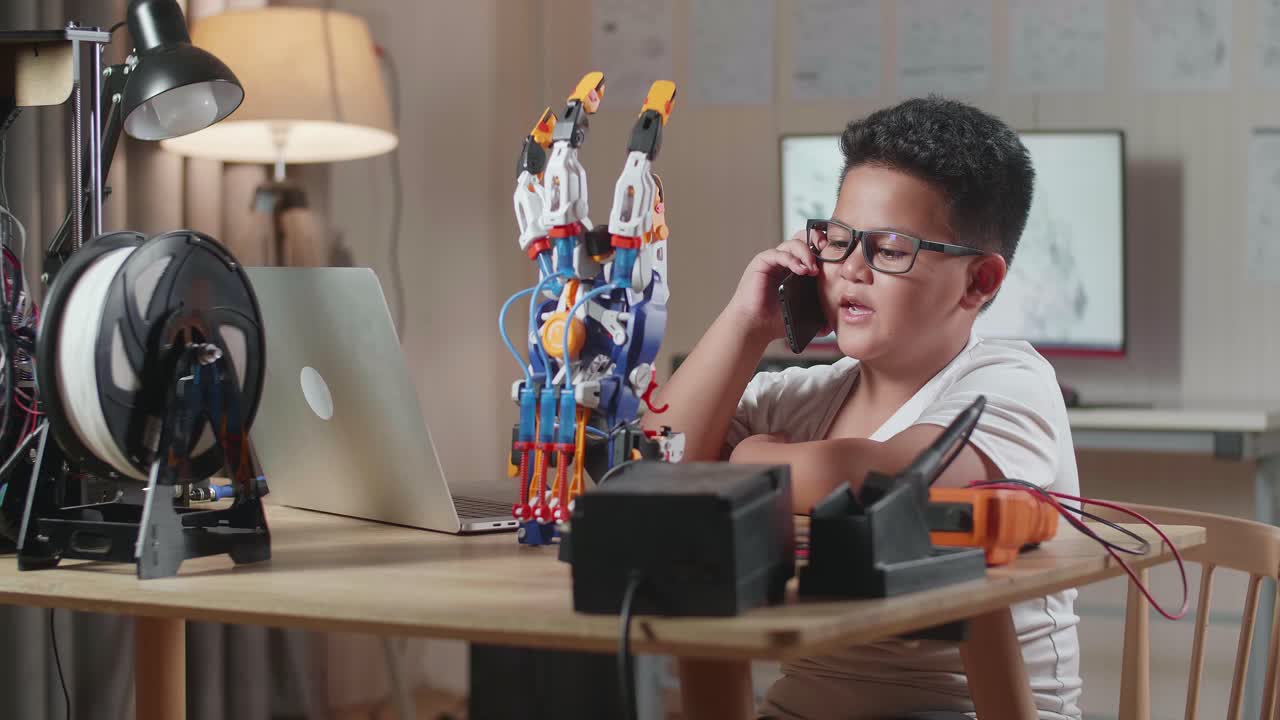 귀여운 아시아 소년이 집에 있는 노트북에서 사이보그 손에 대해 작업하는 동안 스마트폰으로 이야기하고 있습니다.