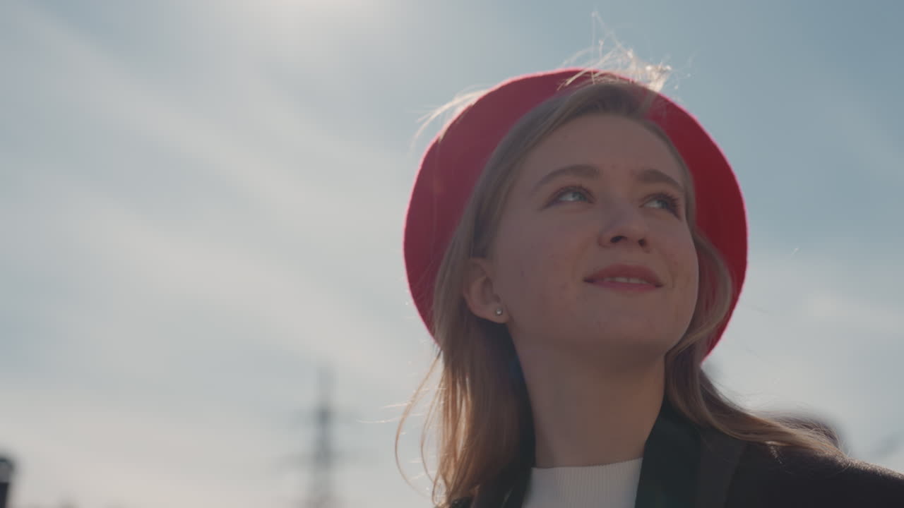 Donna bianca con berretto rosso che guarda il cielo su una strada cittadina illuminata dal sole, indossando cappotto e sciarpa, sorriso sottile e retroilluminazione morbida, capelli biondi e lentiggini leggere, un vecchio edificio rosso sfocato sullo sfondo, stagionale.
