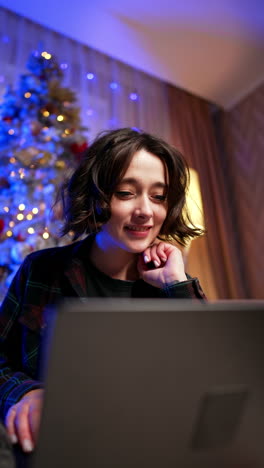 happy smiling woman looks at her laptop. girl leans to the screen and sits pensively. christmas tree at backdrop. vertical video.